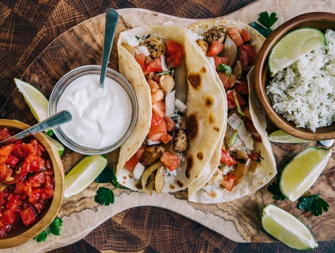 An olive board filled with delicious mexican fiesta of food tacos and burritos with cheese, onions, chicken, pico de gallo, cilantro lime rice and sour cream