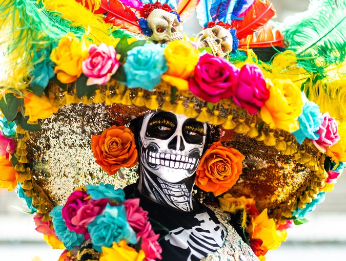 Person sminkad som ett skelett bärandes stor hatt med blommor. Fotografi.
