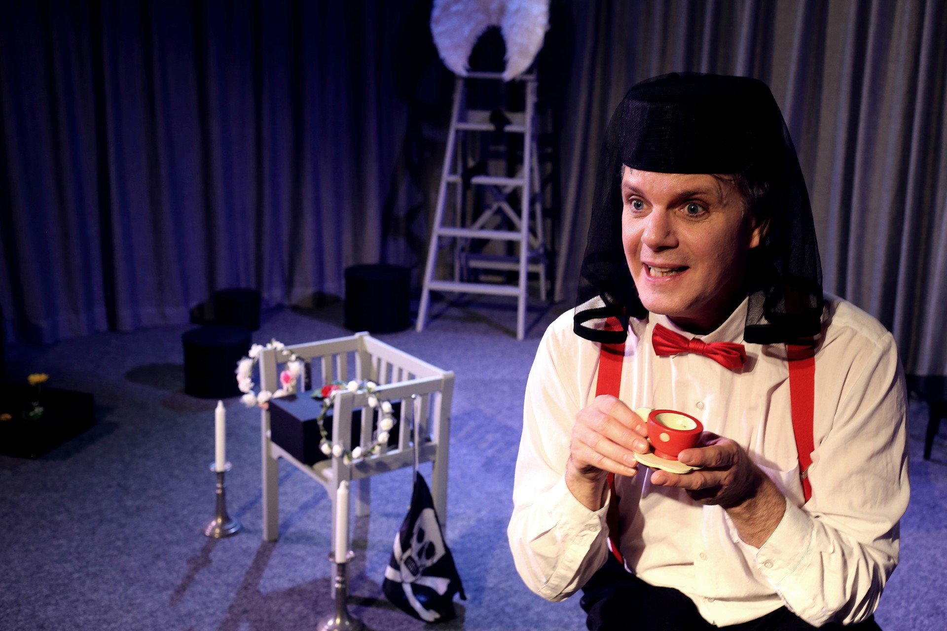 Bilden visar en person som sitter i förgrunden, iklädd en vit skjorta, röda hängslen, en röd fluga och en svart slöja över huvudet. Karaktären håller i en liten röd kopp med vita prickar och ett matchande fat.  I bakgrunden finns en stege och en liten vit stol som liknar en spjälsäng prydd med kransar av vita blommor. Ett svart tyg med en dödskalle hänger på ena sidan av stolen. Framför stolen står två stearinjus.  Bakom spjälsängen syns en stege med ett par stora vita vingar fästa på toppen. Det finns också en gul blomma på golvet.