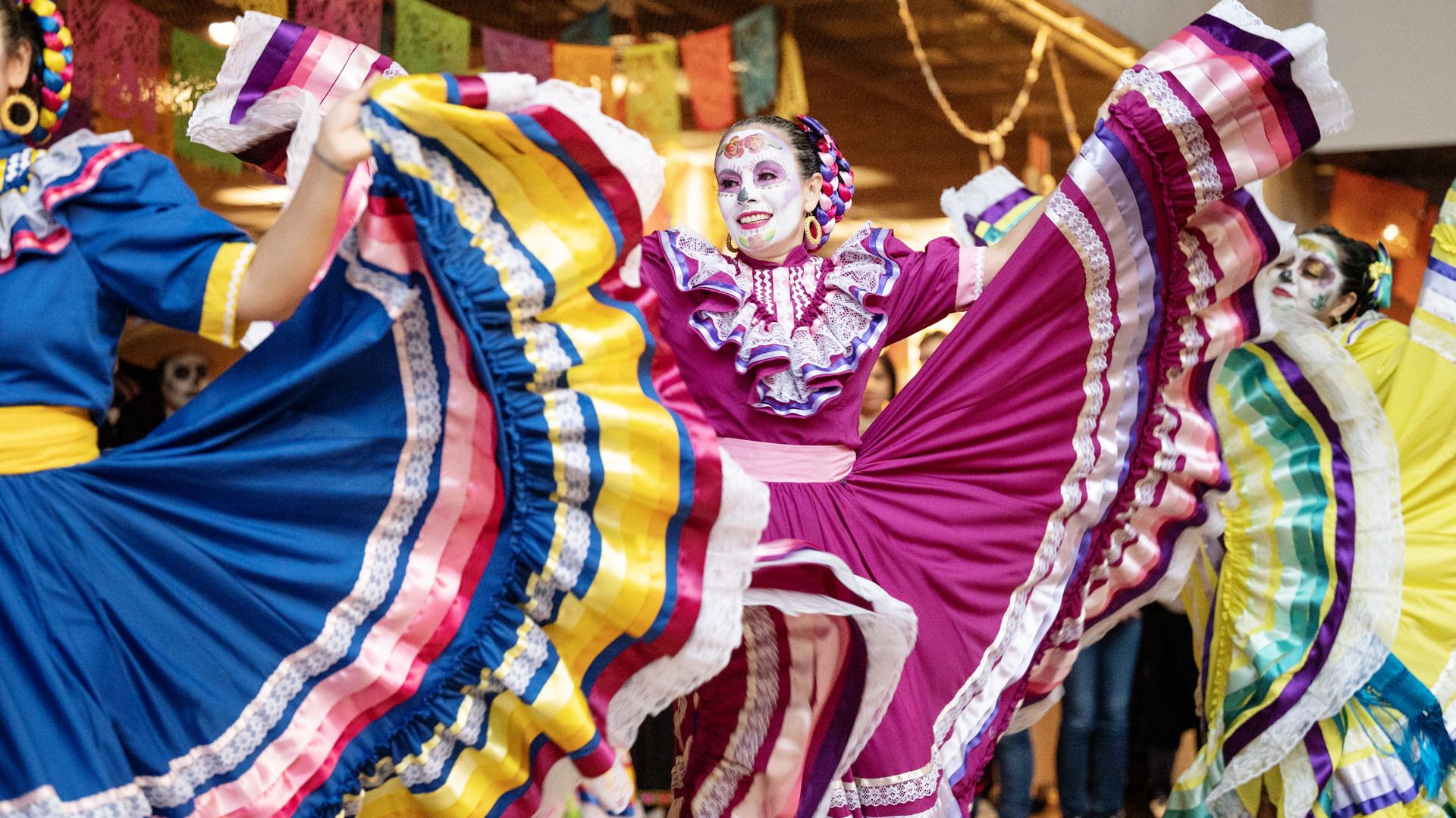 Dansgruppen Mexicas uppträder i färgstarka traditionella dräkter med vida kjolar som hålls ut i rörelse. Flera dansare bär ansiktsmålning inspirerad av Día de los Muertos, och färgglada pappersdekorationer syns i bakgrunden. Bilden förmedlar en livfull scen med fokus på dans, kultur och rörelse.